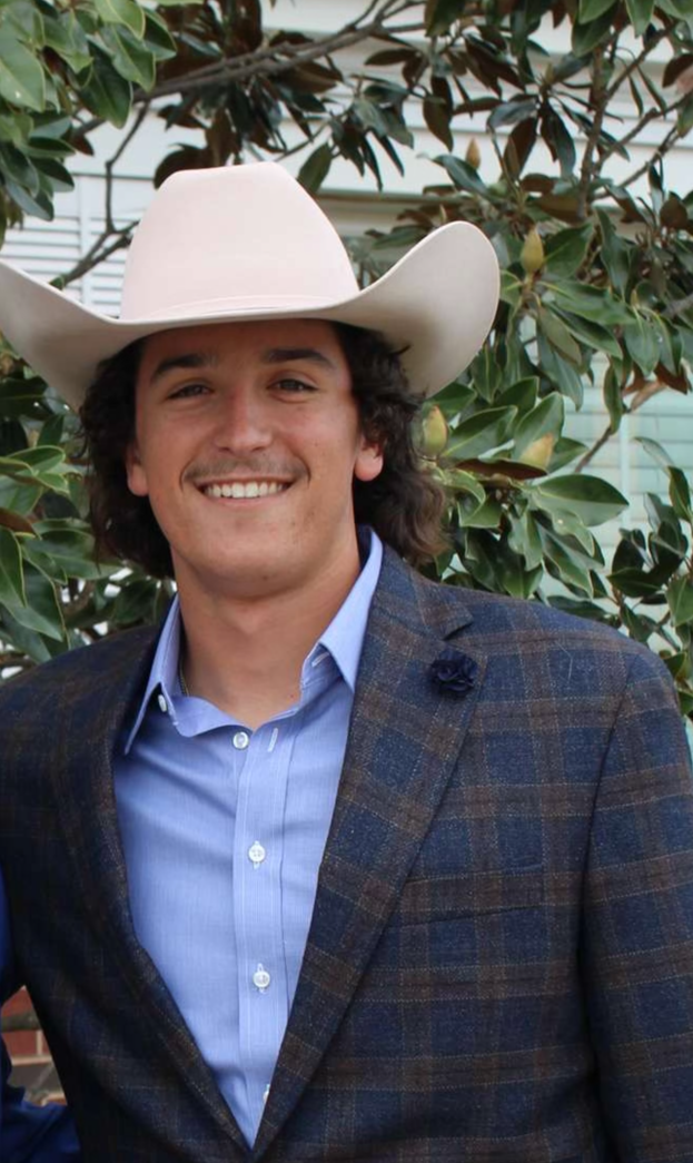 A man wearing a cowboy hat and a suit jacket is standing in front of a tree.