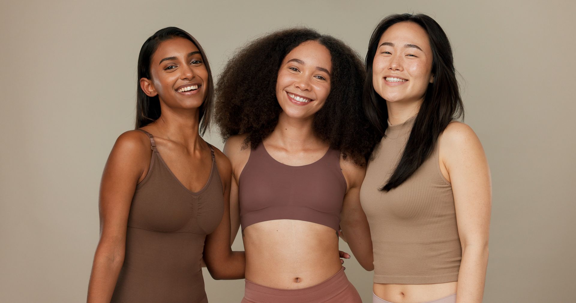 Three diverse women smiling and embracing, wearing neutral-toned activewear against a beige backdrop.