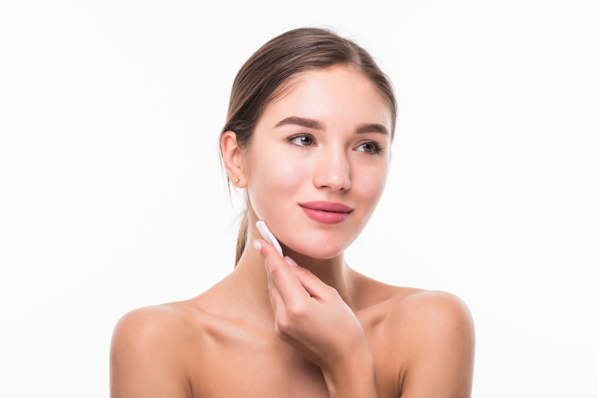 Woman cleaning her face with a white cotton pad, bare shoulders, neutral expression, white background.