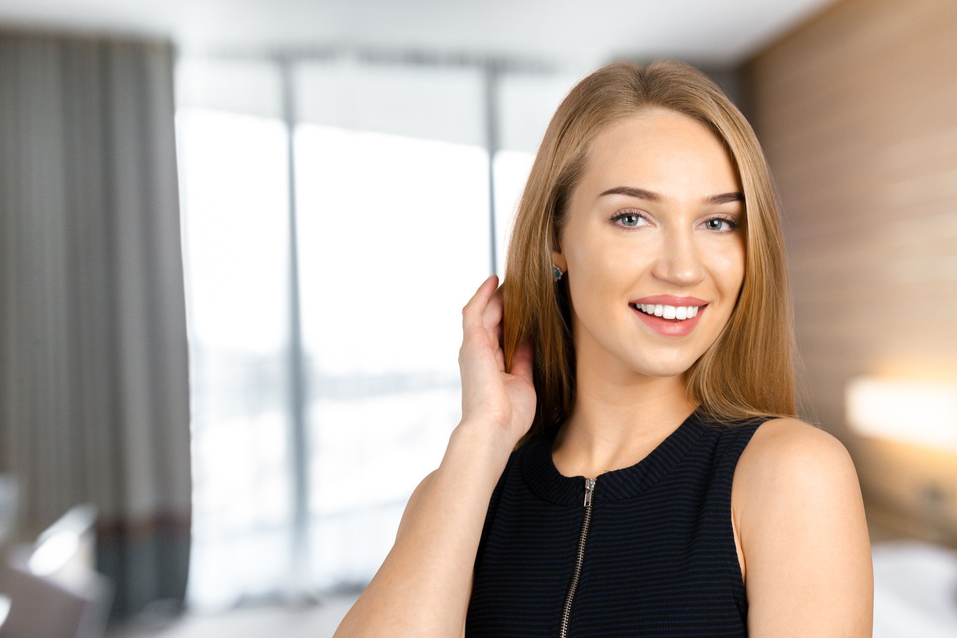 A woman in a black dress is smiling and touching her hair in a bedroom.