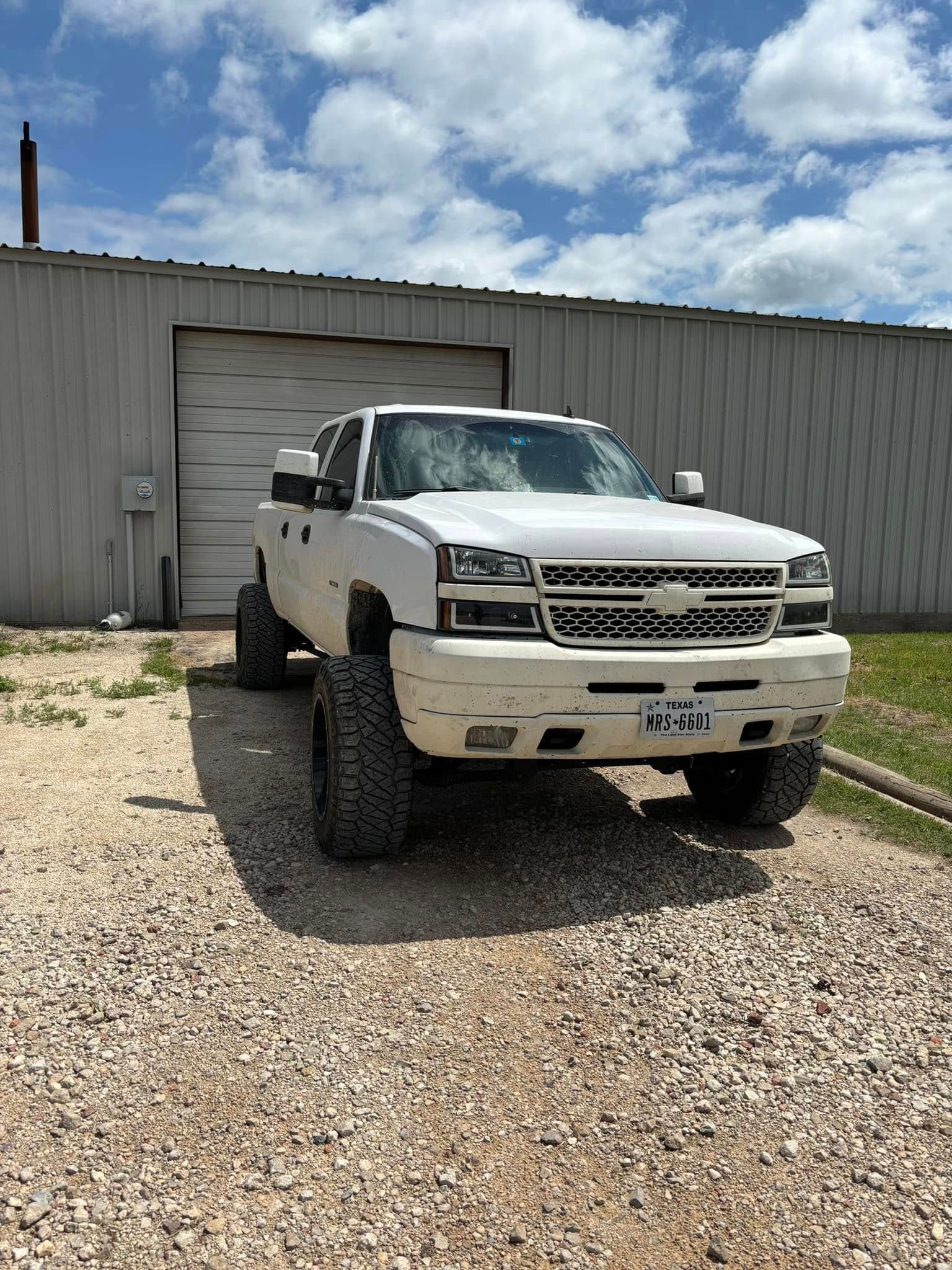 White lifted pickup truck parked in front of a gray industrial building on gravel.