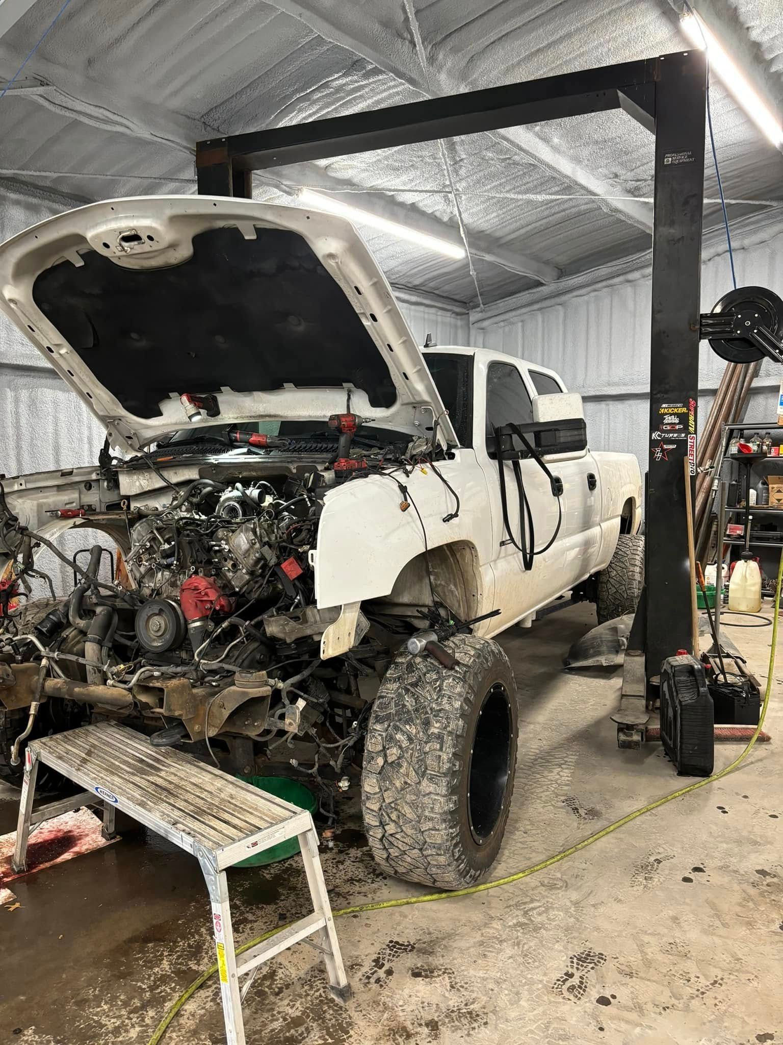 White truck on a lift, engine exposed. Garage setting with tools and open hood.