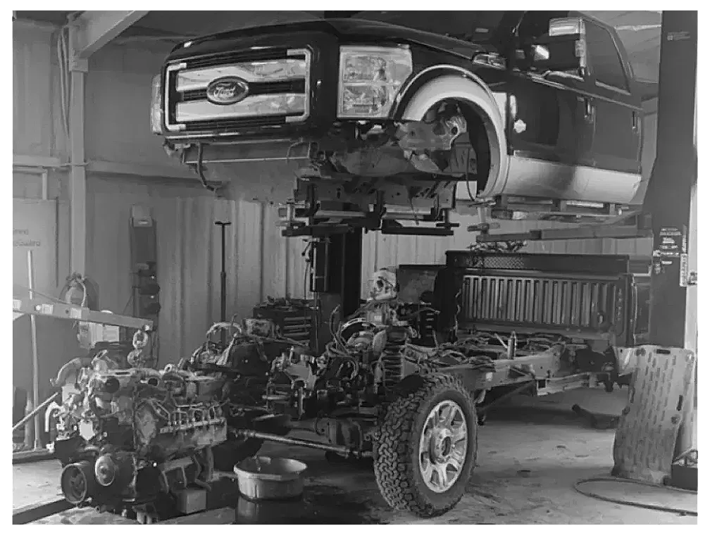 Black and white photo of a pickup truck being worked on in a garage. The cab of the truck is elevated above the frame.