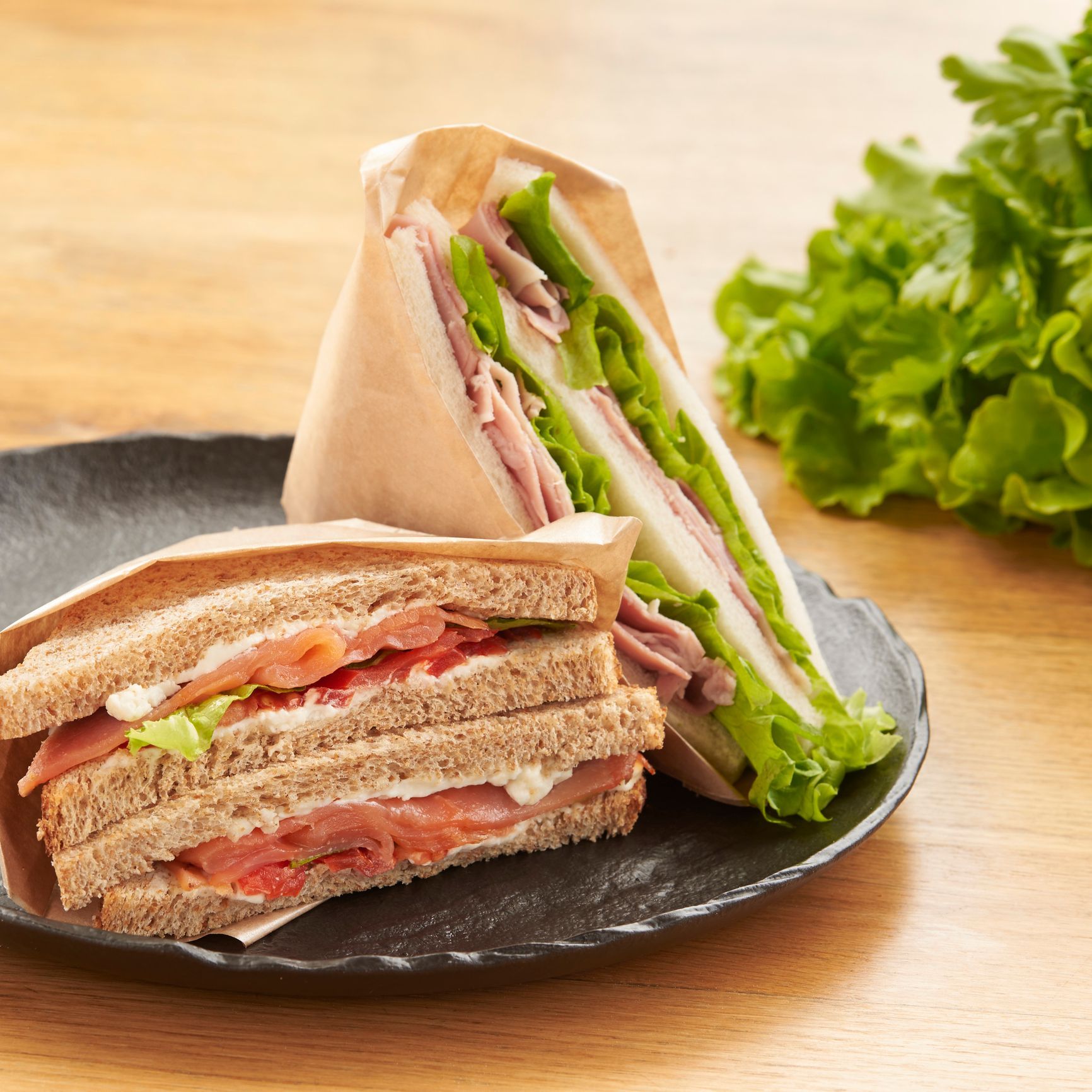 Due panini su un piatto scuro, avvolti nella carta; ripieno di lattuga e carne, con un rametto di lattuga sullo sfondo.