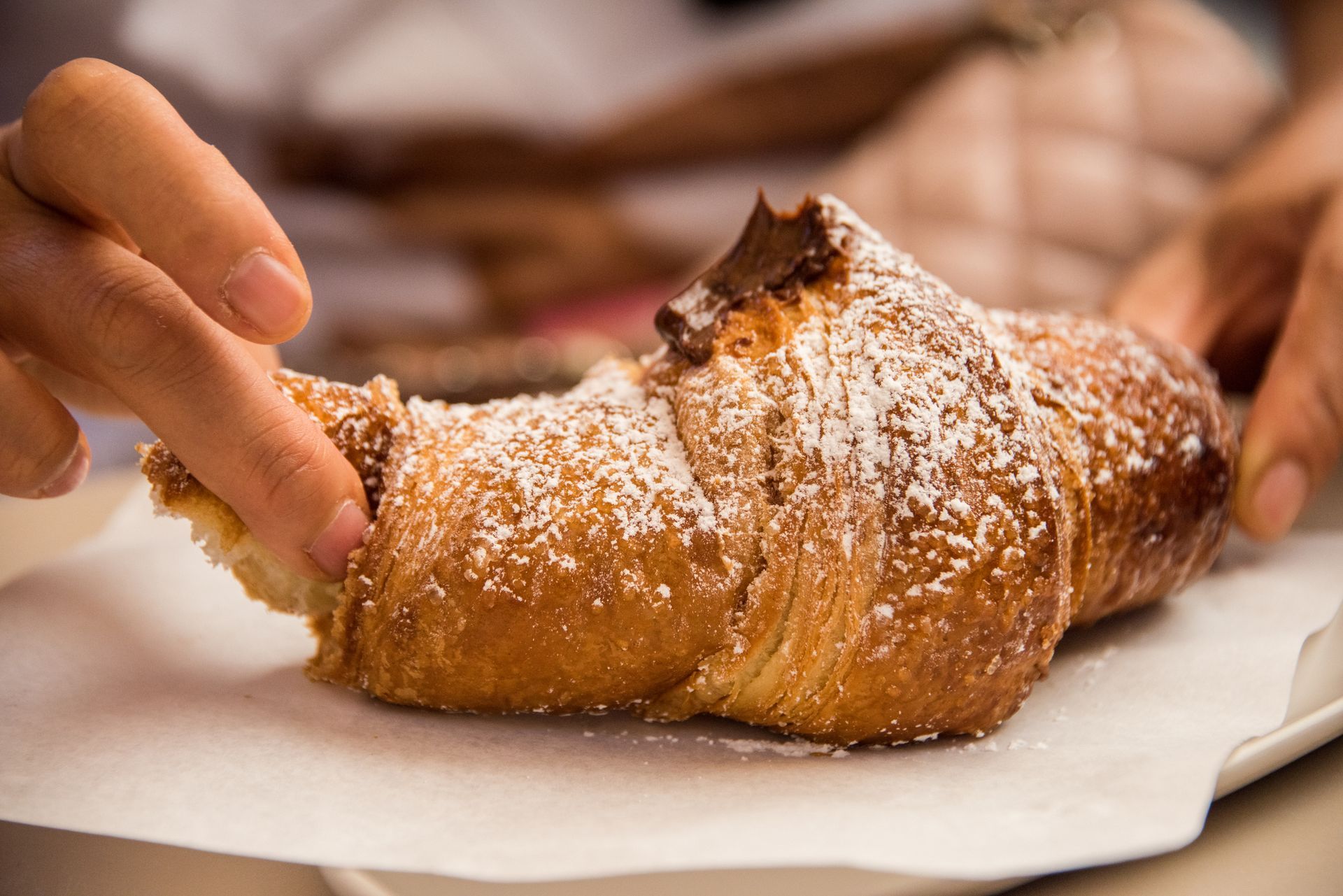 Mano di una persona che spezza un croissant spolverato con zucchero a velo su un foglio di carta bianca.