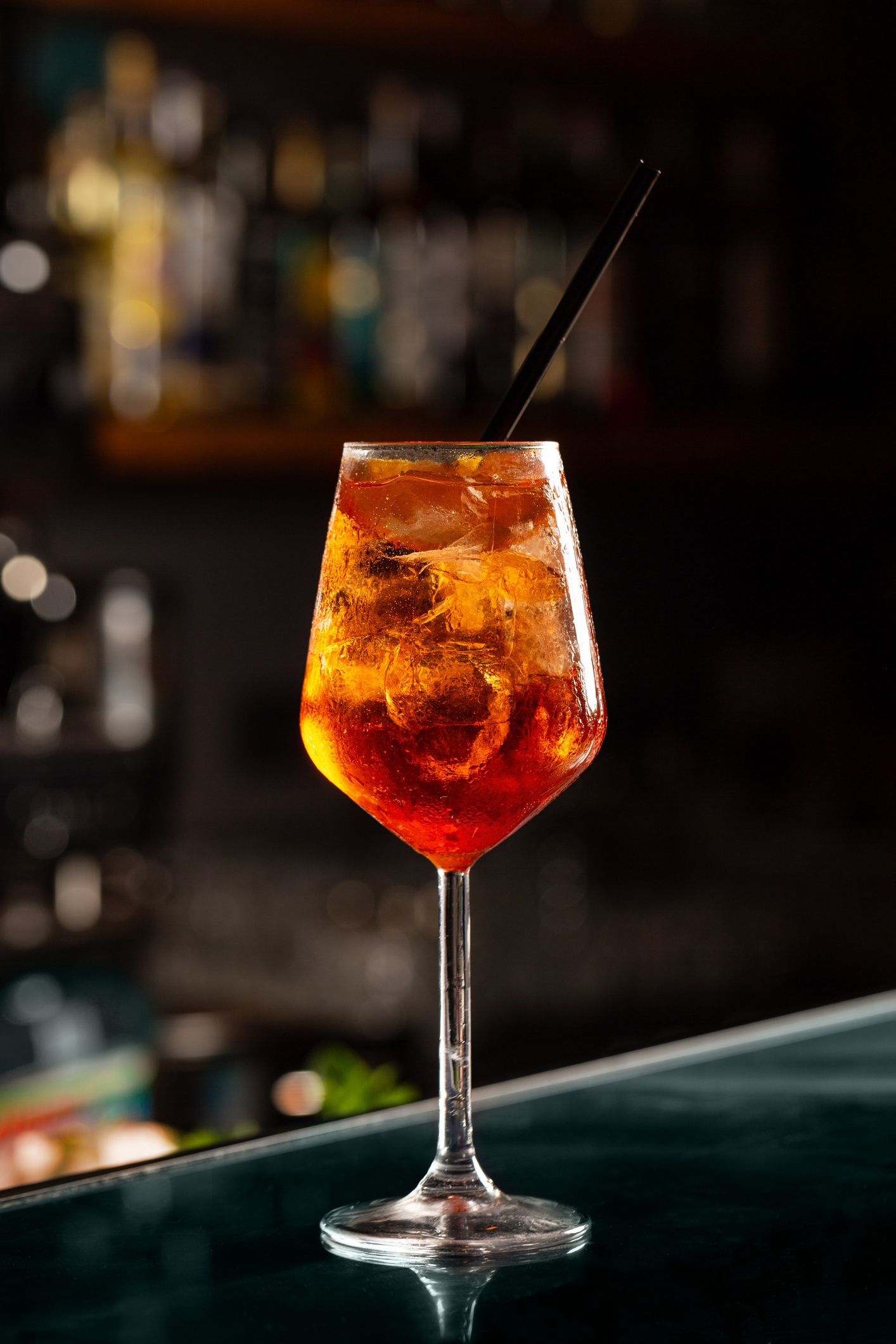 Cocktail alto in un bicchiere a stelo, liquido ambrato, ghiaccio, una cannuccia scura e uno sfondo sfocato del bar.