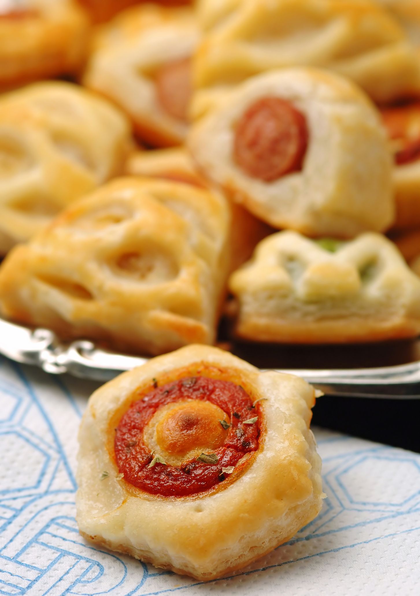 Primo piano di antipasti assortiti di pasta sfoglia, tra cui involtini di salsiccia e spirali di pomodoro, su un vassoio.