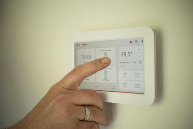A hand touches a touchscreen thermostat mounted on a beige wall, adjusting the temperature.