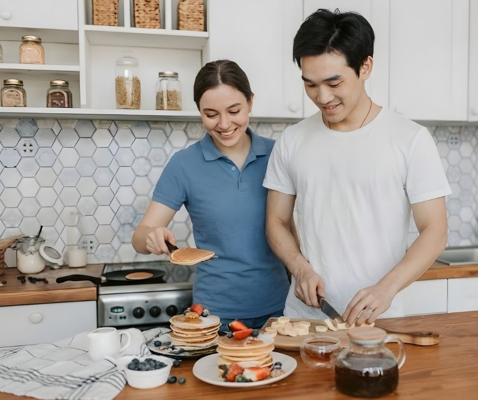 A female support worker and a young male NDIS participant smiling while preparing a pancake breakfast together in a modern home kitchen. This visualizes practical daily living assistance with tasks like meal preparation, helping the participant regain autonomy in a safe, stable home environment.