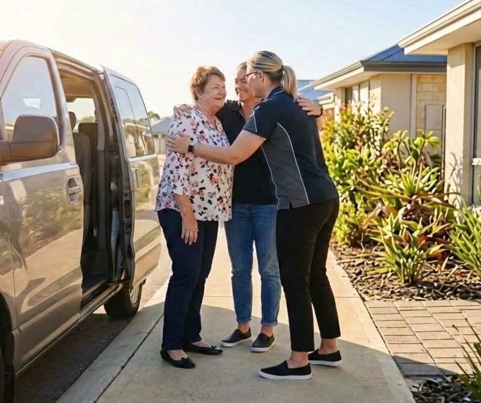 A smiling NDIS participant being greeted warmly by support workers after arriving in an accessible transport vehicle on a sun-drenched Perth residential street. This represents the final stage of a successful transition of care, highlighting the joy of reconnecting with friends and family and returning to a safe, stable home within the community.