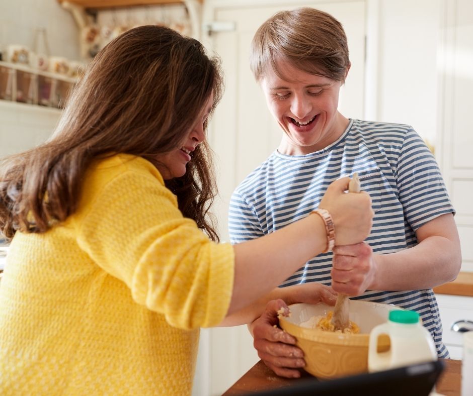 An NDIS supporter and participant preparing a meal together in a warm, domestic kitchen, representing the stable, long-term home environment provided by Supported Independent Living (SIL) to facilitate a permanent return to the community and reduce the risk of hospital readmission.