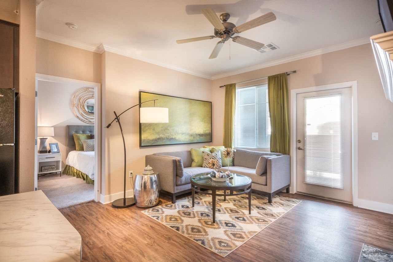 Living room in an apartment with a gray sofa, glass coffee table, area rug, and doorway to a bedroom.