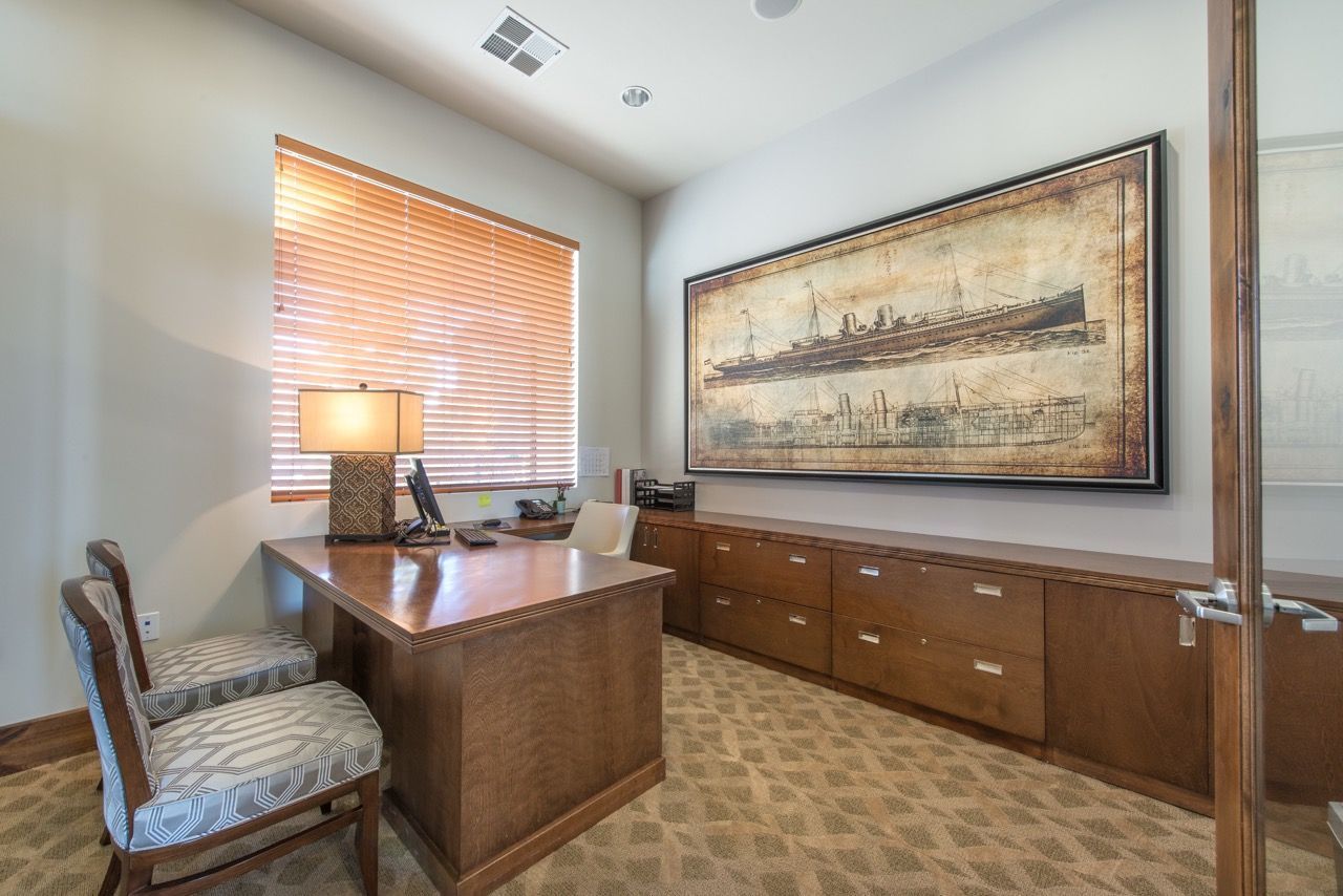 Office interior with a wooden desk, two patterned chairs, and a large framed ship artwork.