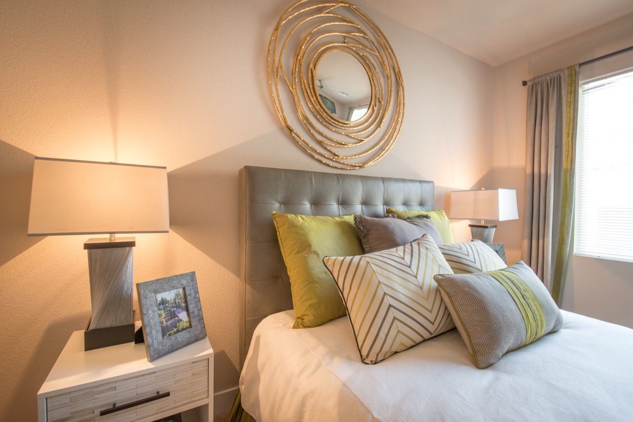 Bedroom in a modern apartment with an upholstered headboard, decorative pillows, bedside lamps, and a round mirror.
