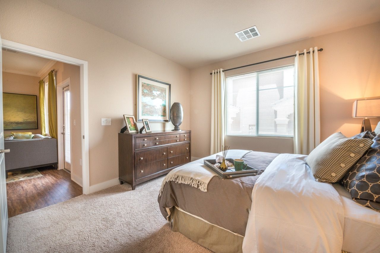 Bedroom in an apartment with a queen bed, dark wood dresser, and a large window with light curtains.