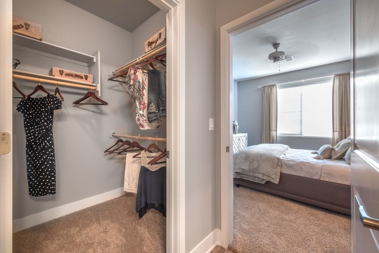 Open walk-in closet with clothes, next to a bedroom with a bed and window.