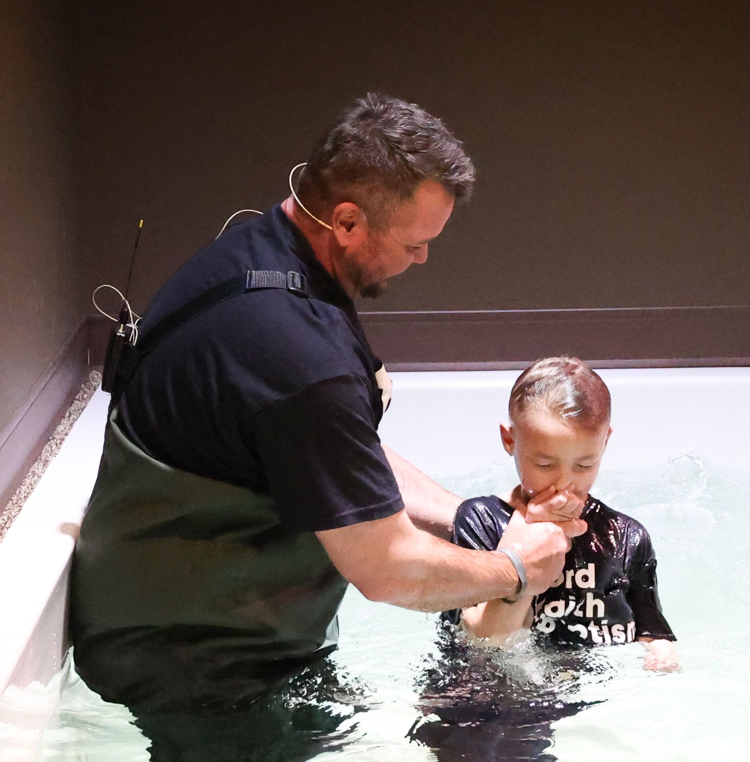 A man is baptising a young boy who is wearing a shirt that says lord loves this