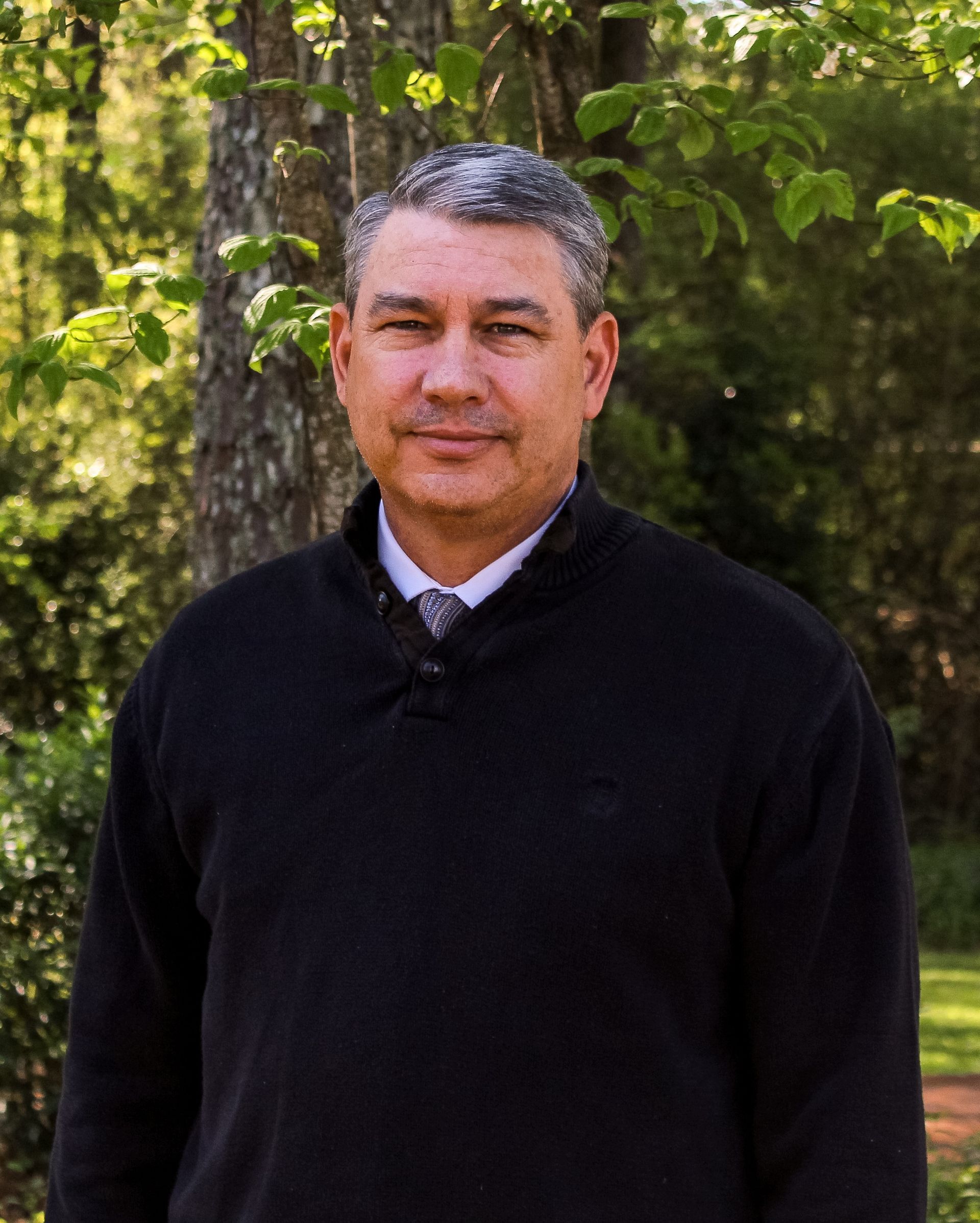 A man in a black sweater and tie is standing in front of a tree.