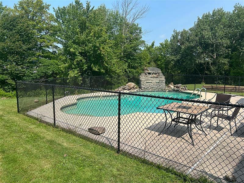 A chain link fence surrounds a swimming pool with a table and chairs.