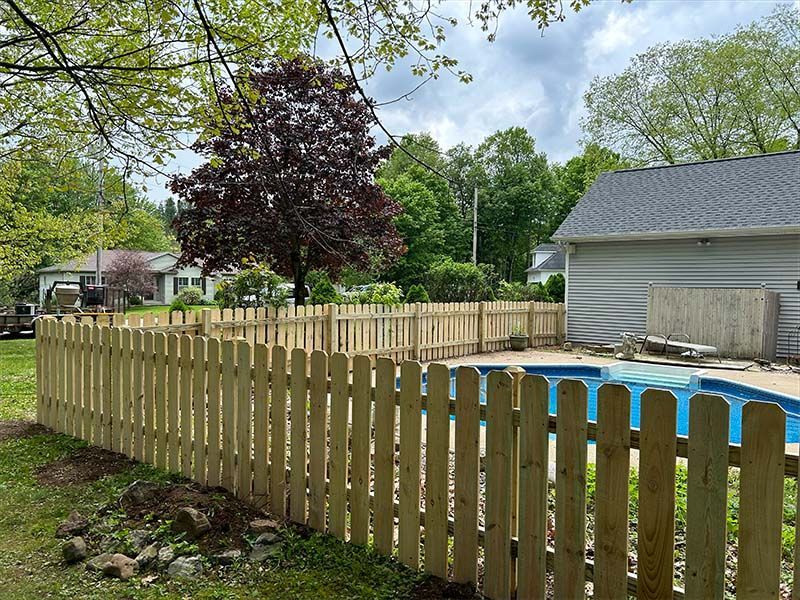 A wooden picket fence surrounds a swimming pool in a backyard.