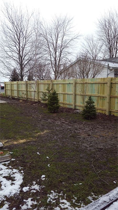 A wooden fence in a backyard with snow on the ground and trees in the background.