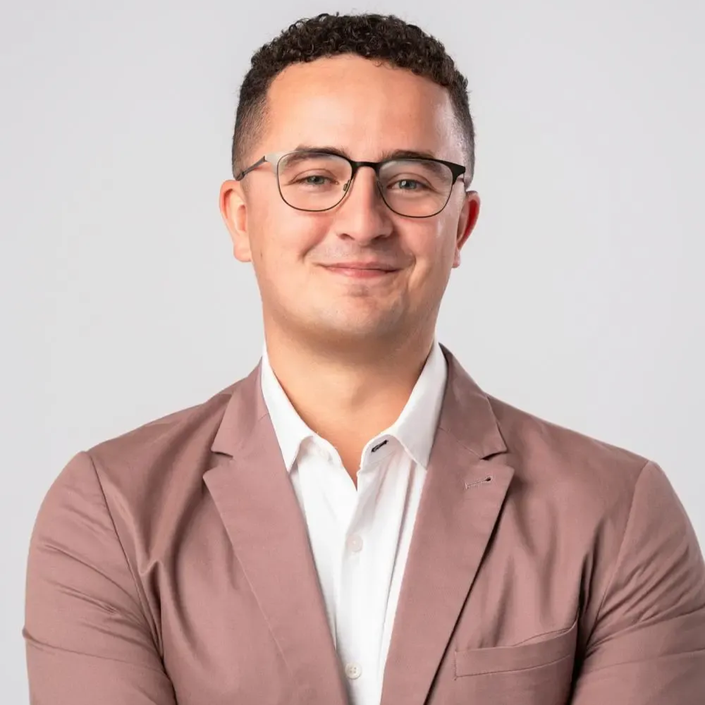 Man with glasses, wearing a pink blazer over a white shirt, smiling.