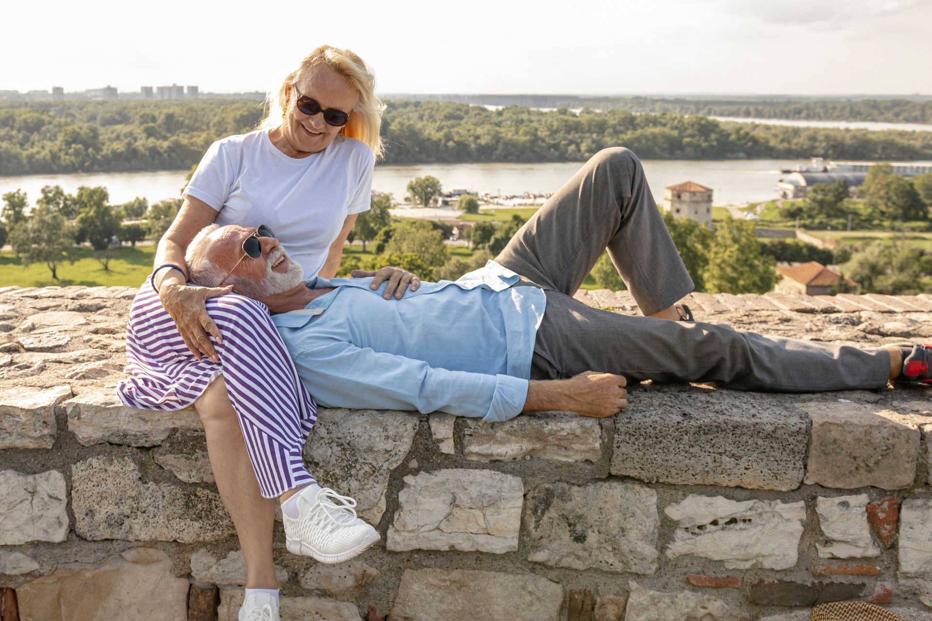 Couple relaxing on a stone wall overlooking a river and city. Man reclines, woman sits beside him.