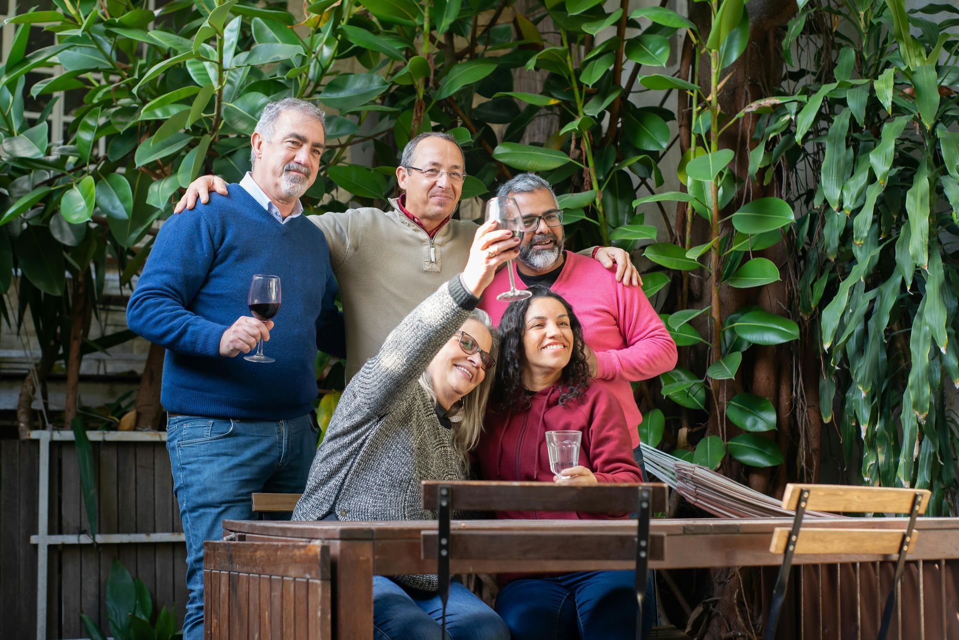 Group of five people smiling, taking a selfie outdoors with wine glasses, lush greenery background.