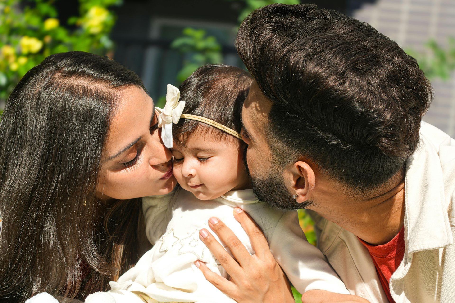 Parents kissing a baby on both cheeks outdoors, sunny day.