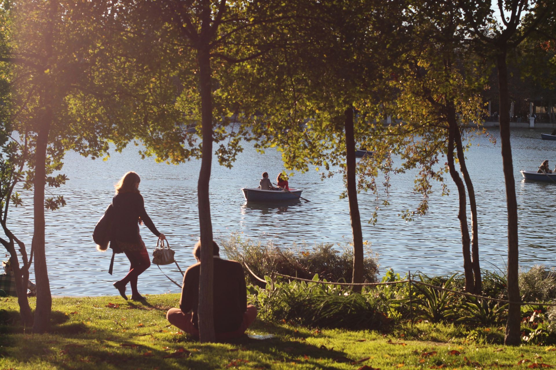 Lakeside scene: person walking with basket, another sitting on grass. Boats on water, trees in sunlight.