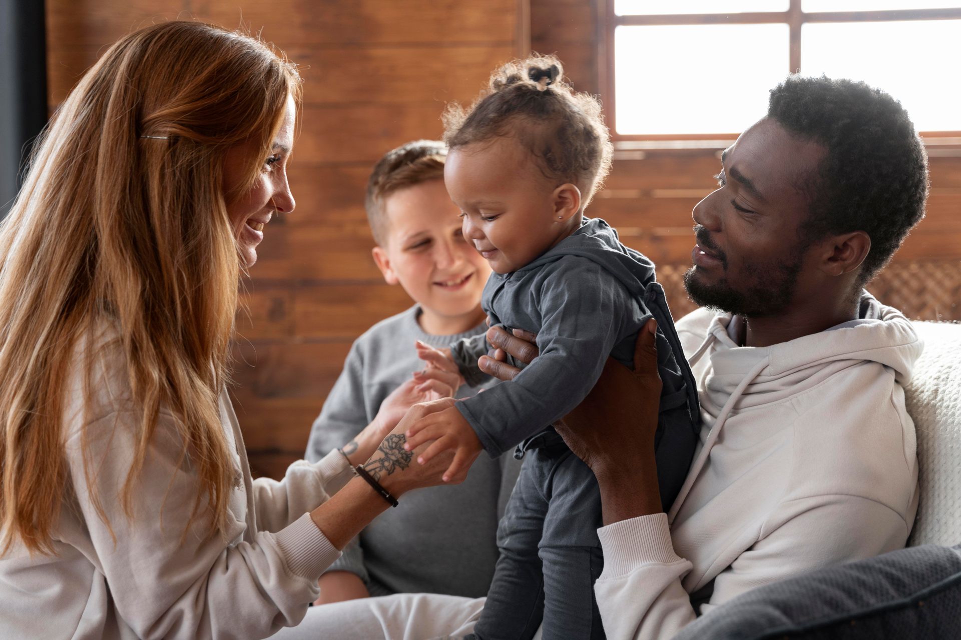 Family indoors, playing with a baby. Parents smile, child looks happy. A sibling watches.