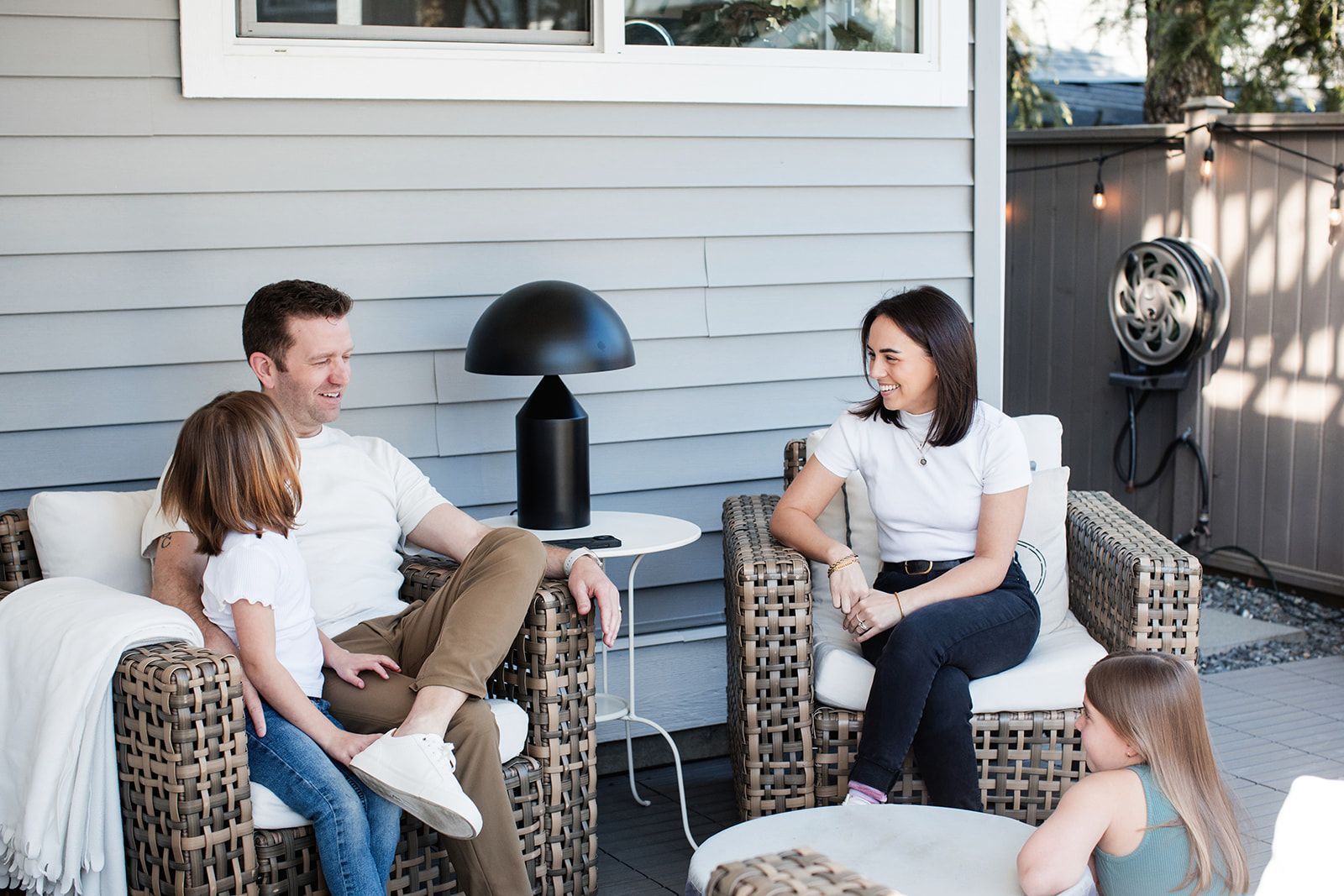 Four people sit on wicker chairs and a bench on a gray patio, chatting beside a house exterior.