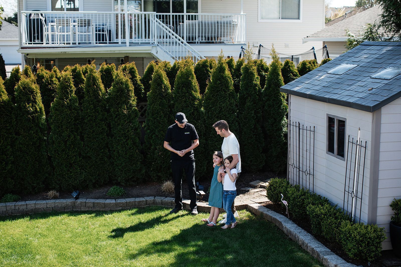 Man in black uniform talking to a family in backyard with a shed and hedge.