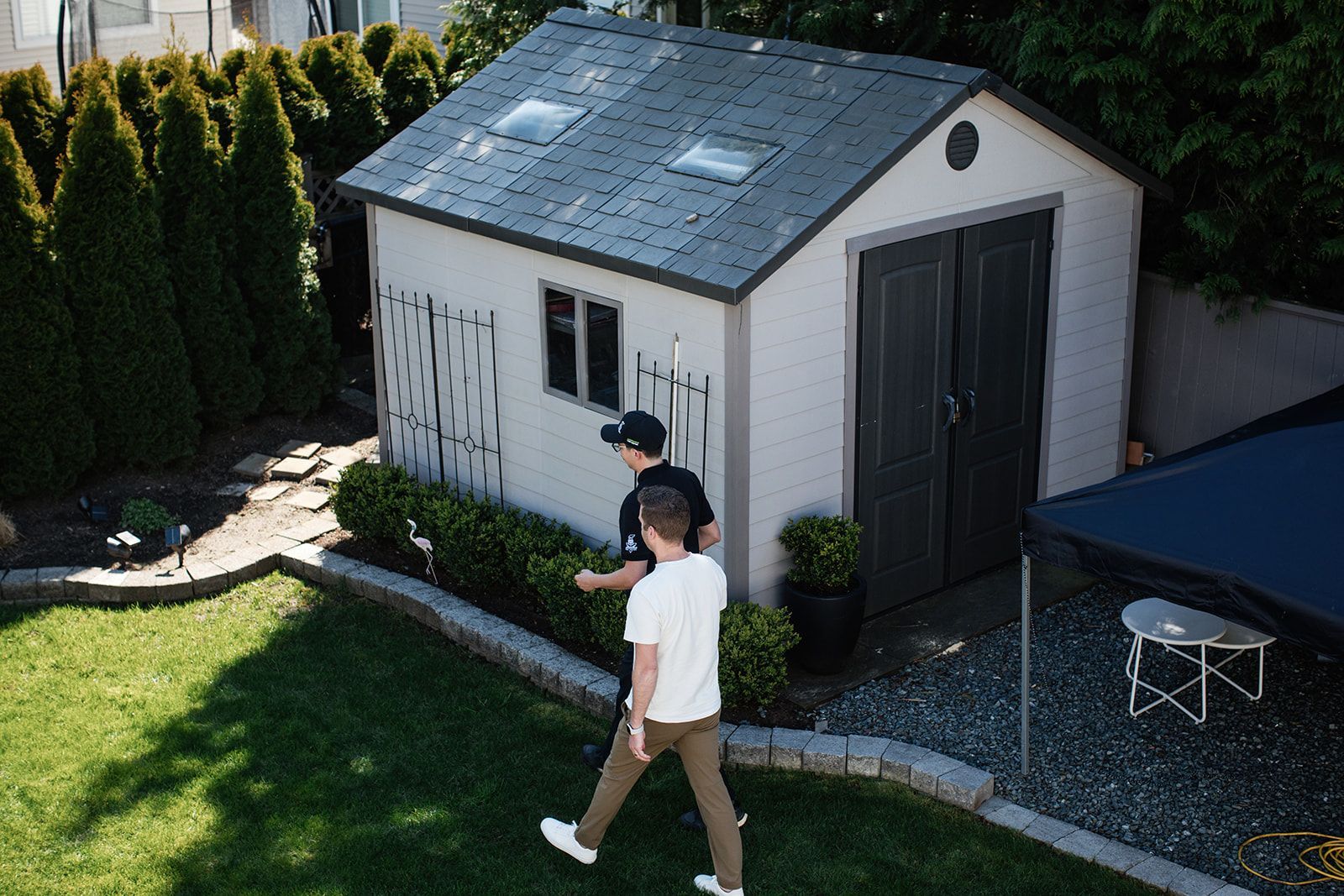 Two people walking beside a white backyard shed with a dark roof and basketball hoop nearby.