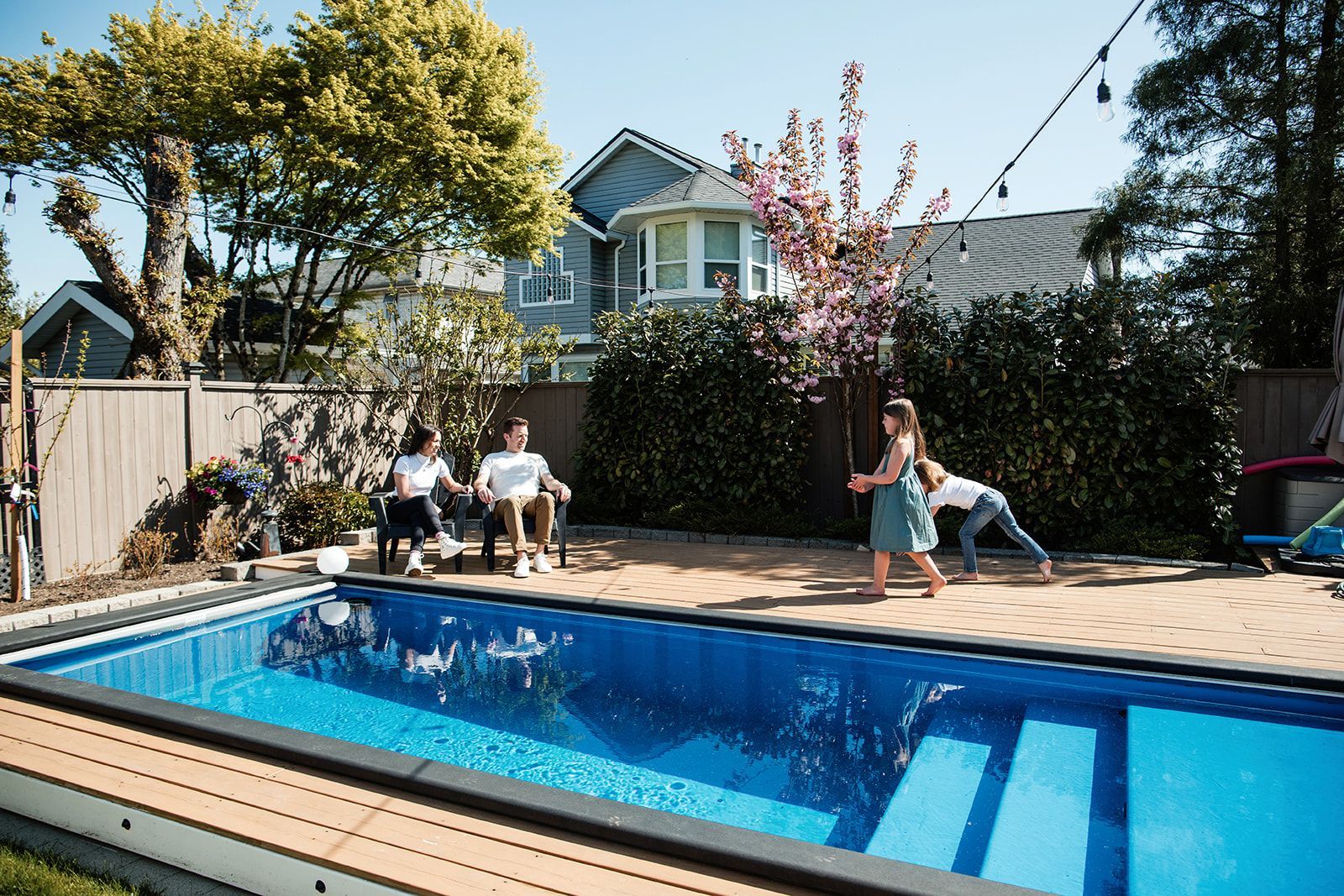 Backyard pool with people lounging on a deck beside a blue house and trees