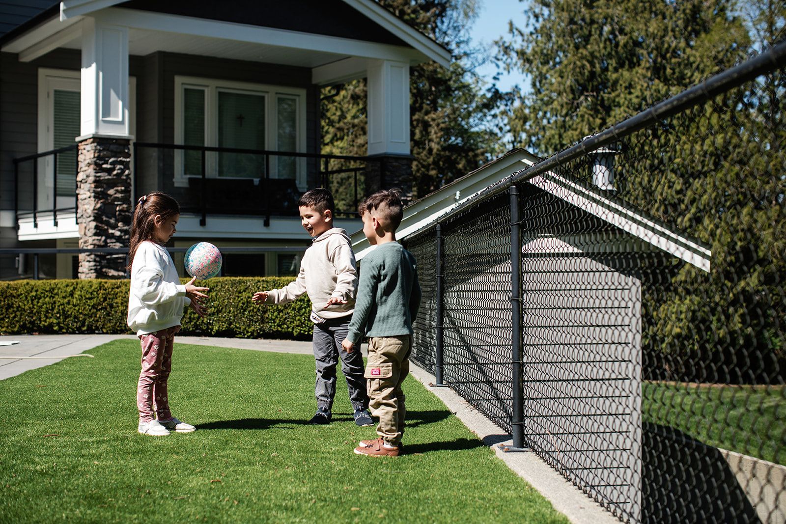 Three children talking on a sunny lawn beside a house and chain-link fence.