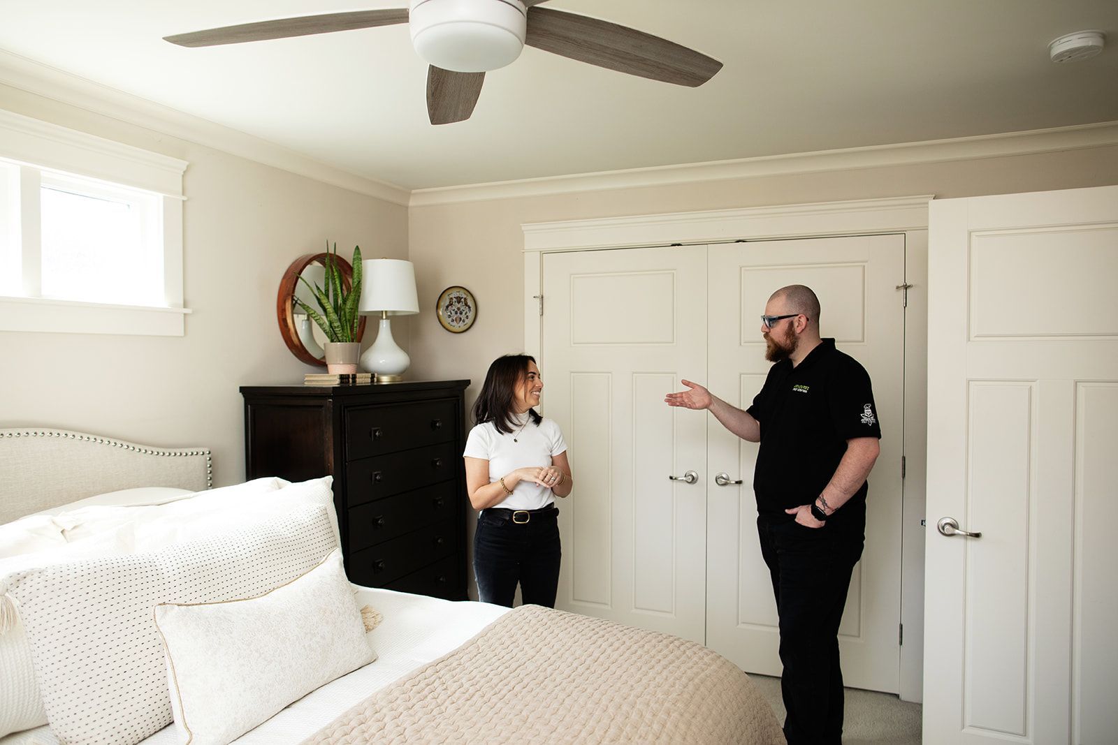 Two people talking in a bright bedroom with a bed, dresser, and ceiling fan.