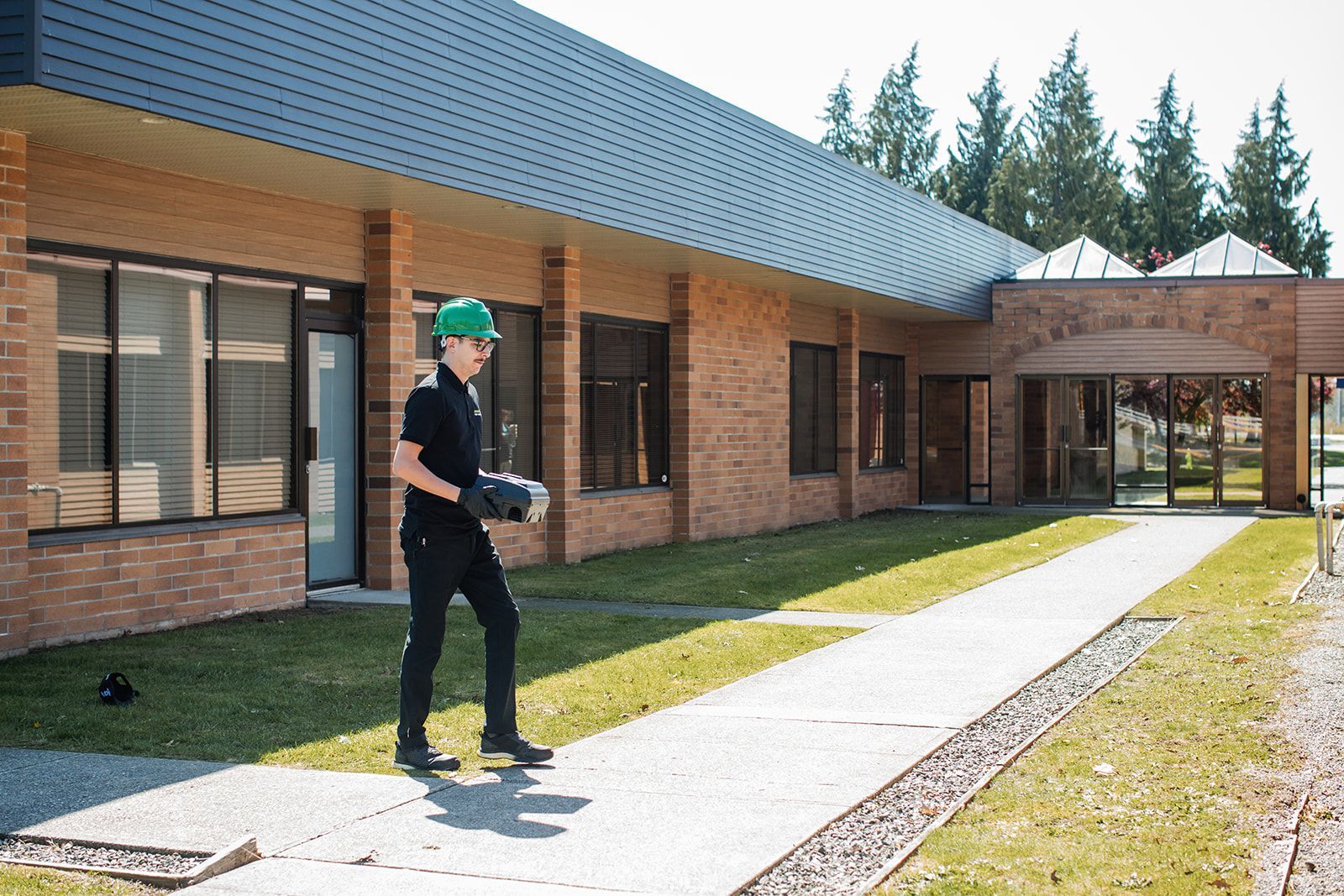 Person in green hard hat standing by a brick building with windows and a covered walkway.