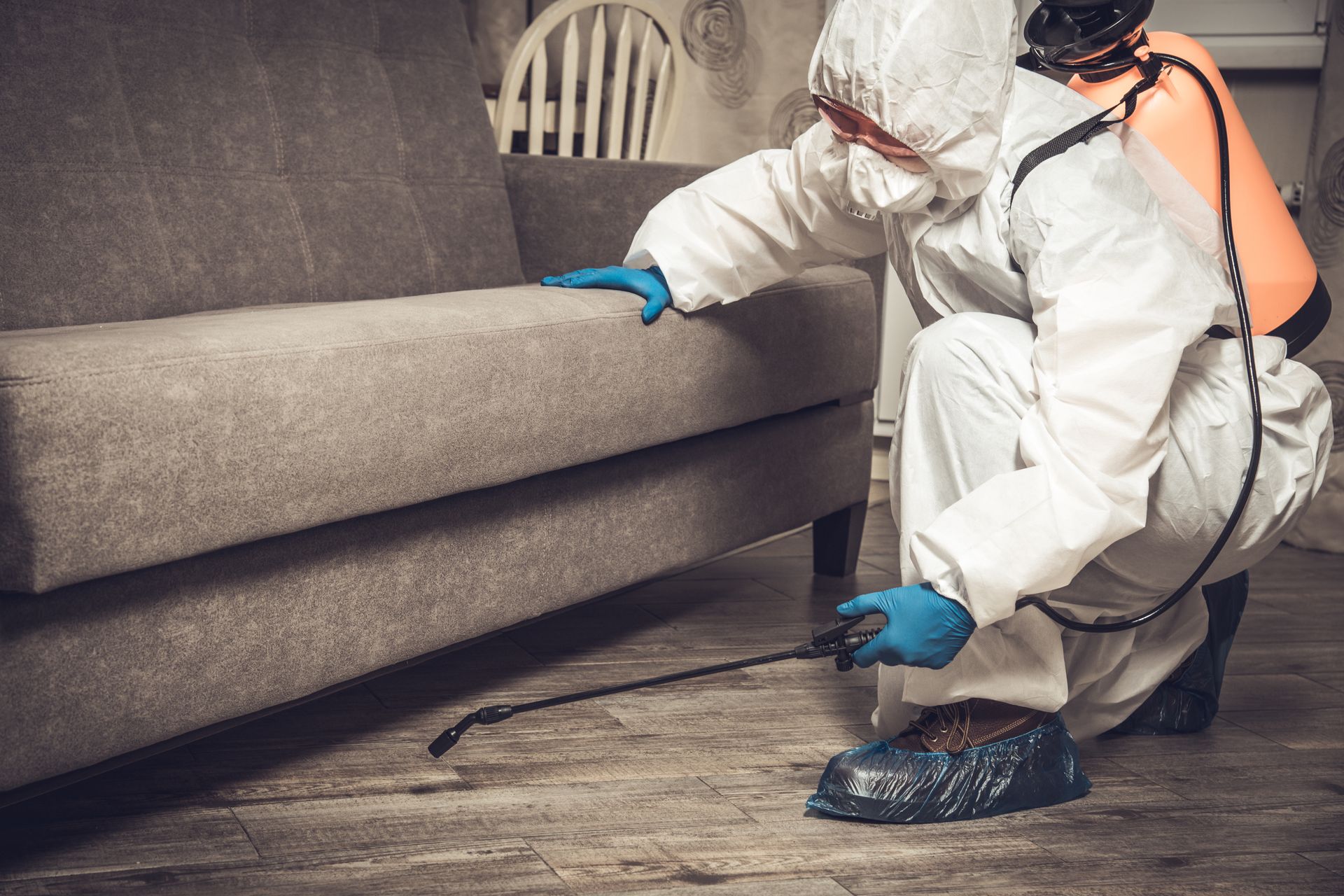 A man in a protective suit is spraying a couch with a sprayer.