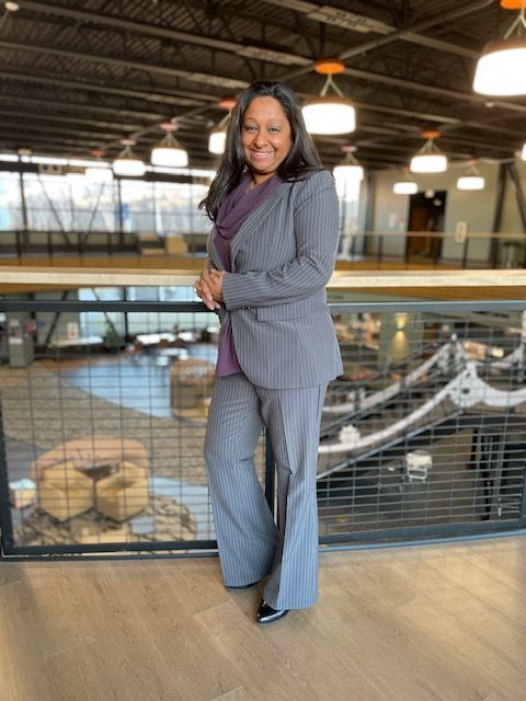 Woman in gray pinstripe suit, standing on a wooden balcony, smiling. Modern, open-concept office.