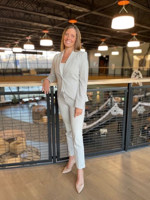 Woman in a light suit smiles, leaning on a railing. Indoor setting with modern decor.