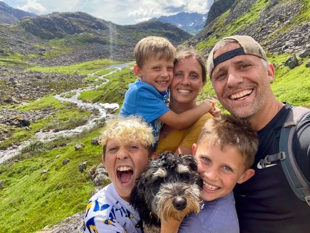 Family of five, including dog, smiling for a selfie outdoors near a mountain stream.