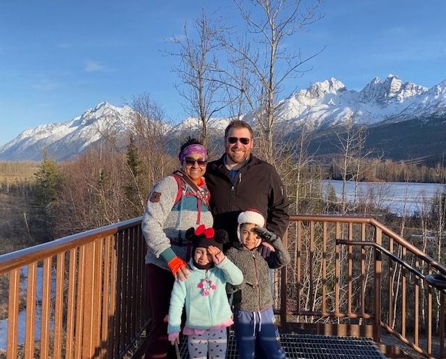 Family poses on a wooden deck with snowy mountains in the background. Blue sky, sunny day.