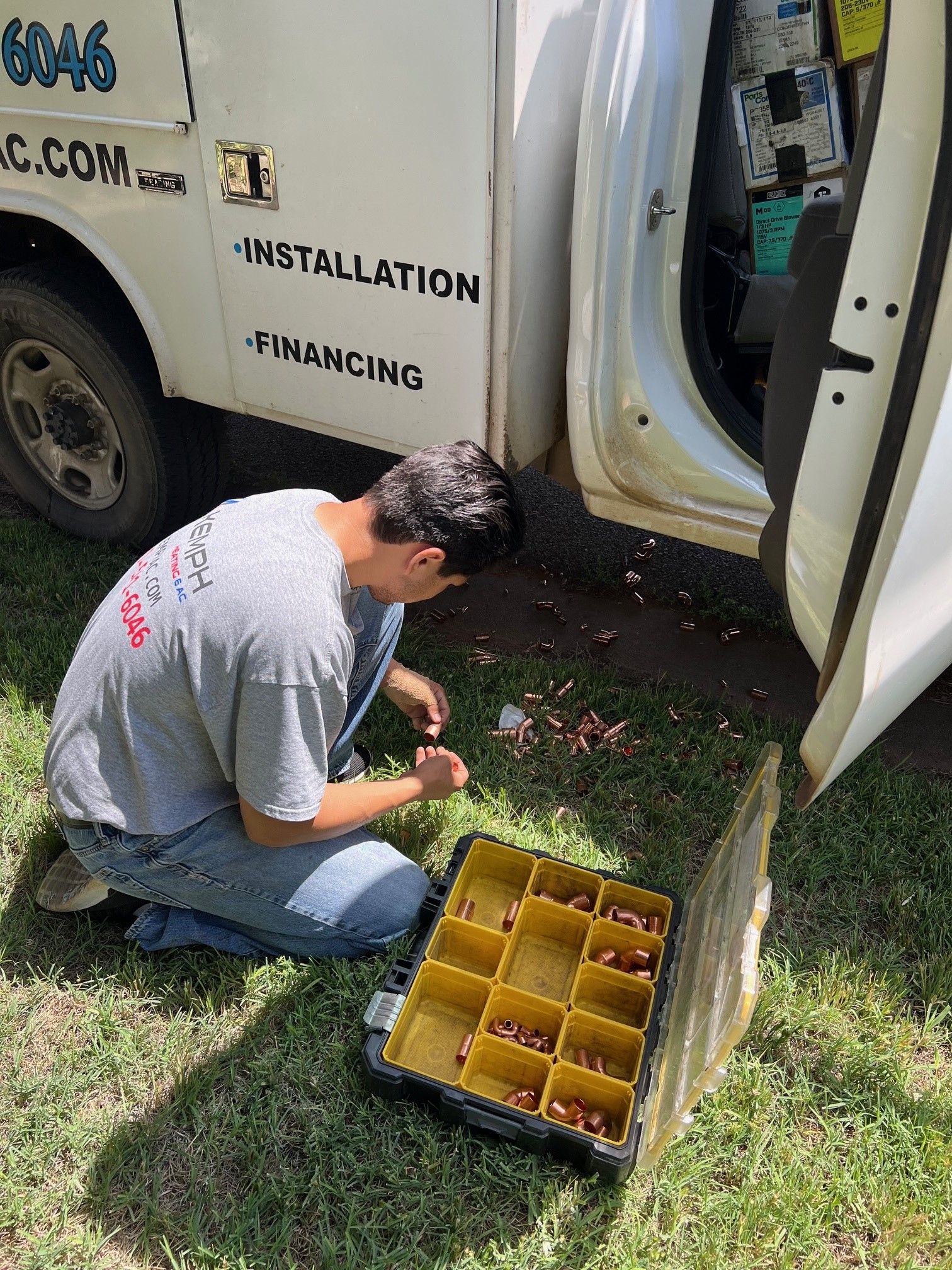 A man kneels by a work van, sorting copper fittings from a tray on the grass.