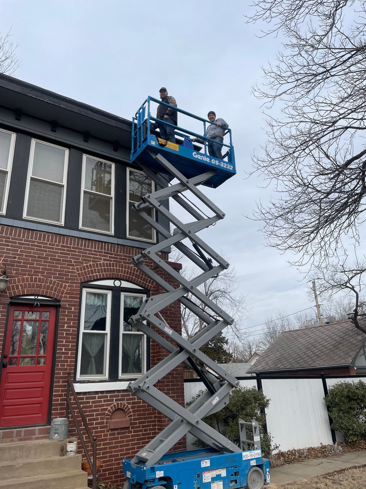 Two people on a blue lift platform working on a building's trim. Brick house, gray sky.