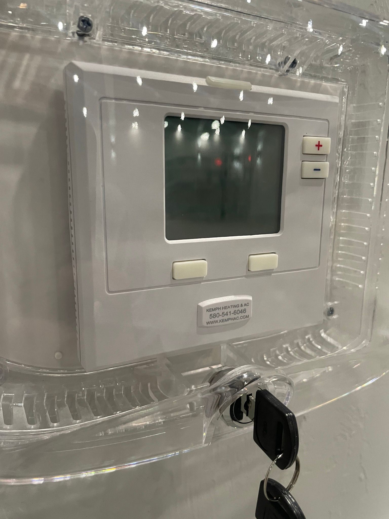 White thermostat with buttons and screen inside a clear acrylic case, keys hanging below.