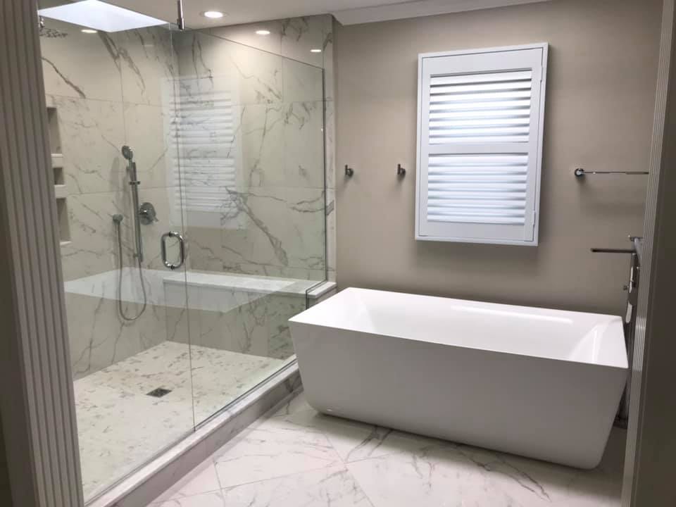 Bathroom with a glass-enclosed shower, freestanding white tub, and shuttered window against a neutral wall.