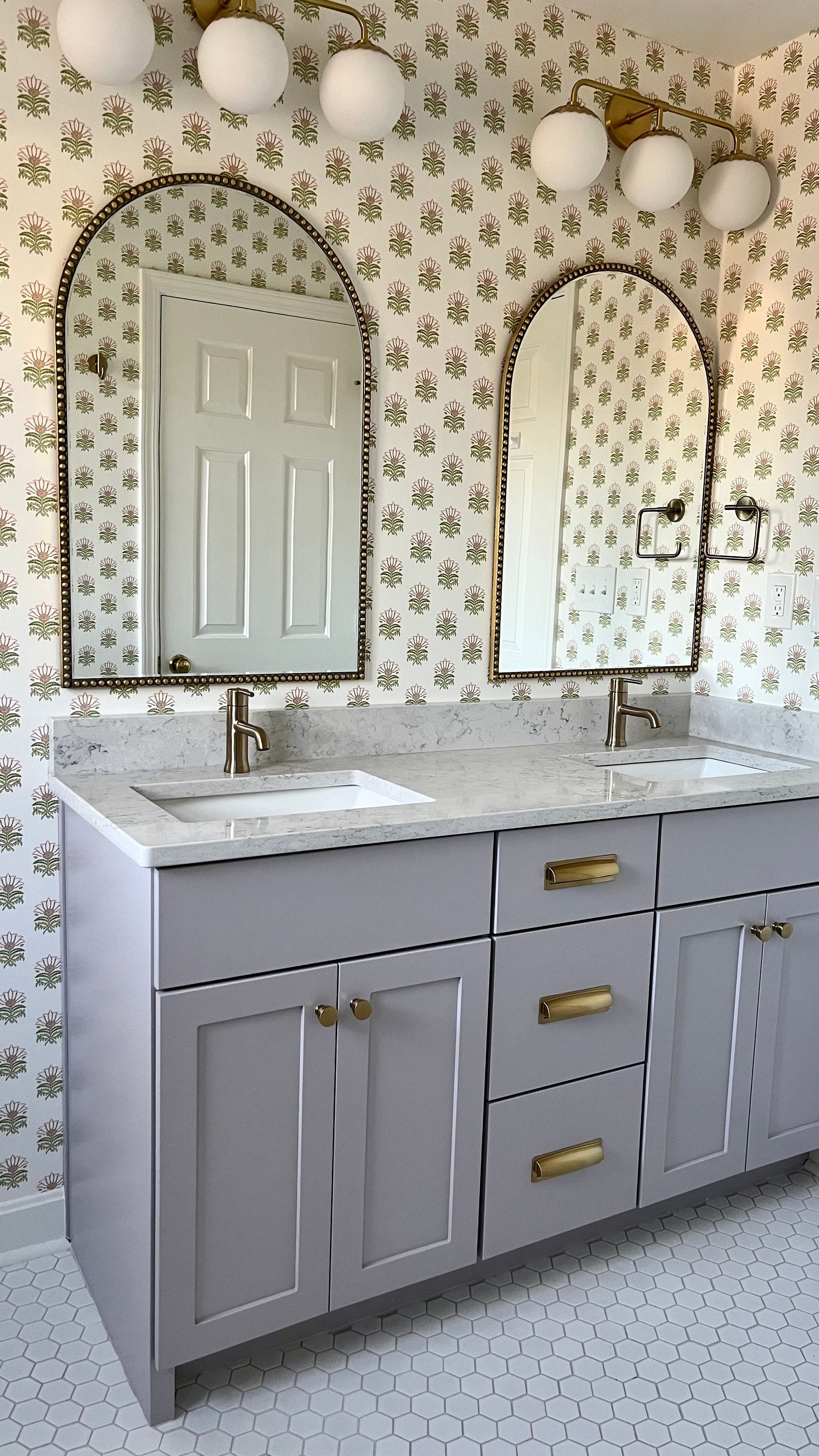 Bathroom with lavender vanity, white countertop, two arched mirrors, floral wallpaper, and gold fixtures.