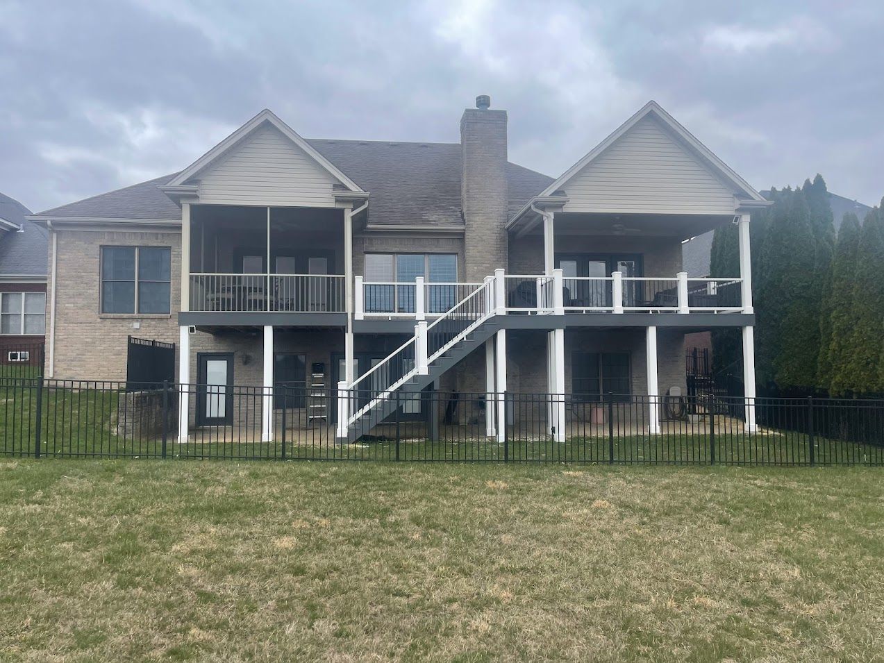Backyard view of a two-story brick house with a deck, stairs, and a black fence. Overcast sky.
