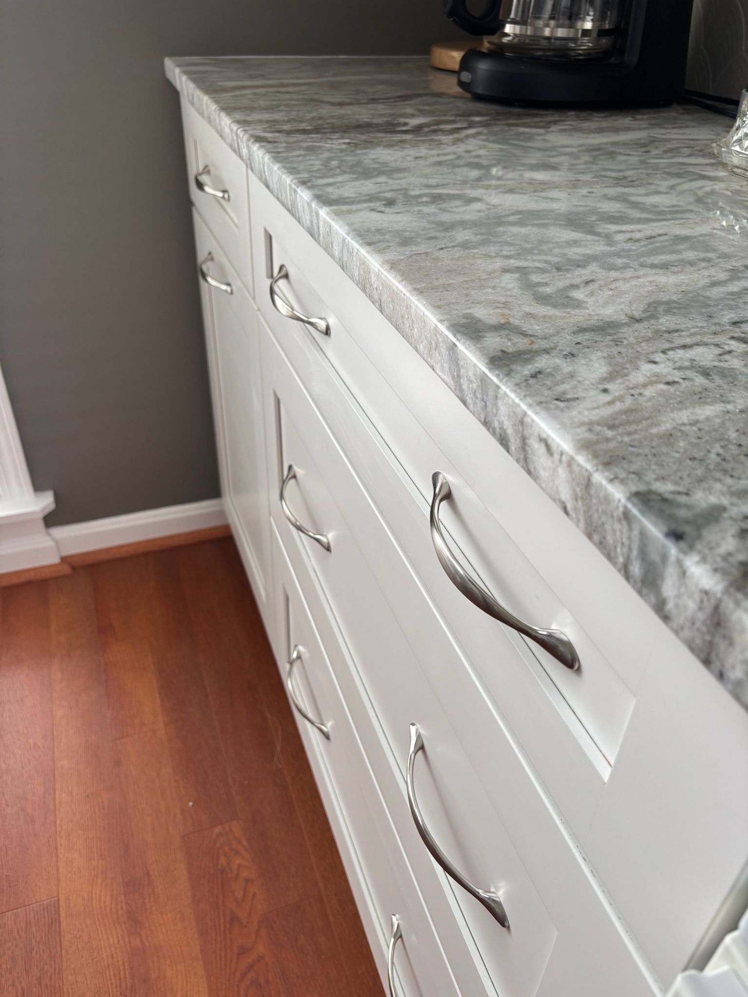 White kitchen cabinets with silver handles under a gray and white countertop.
